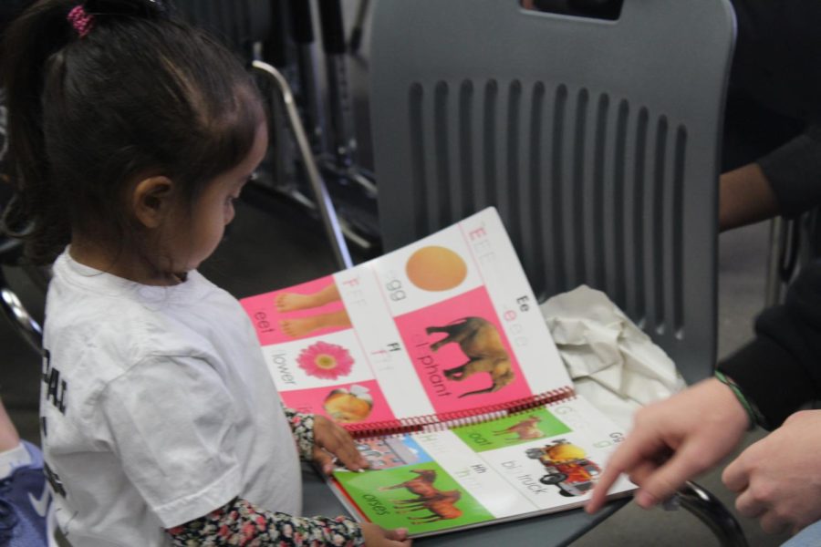 Students help the child read the book they gifted her while others listen to a movie in the background.